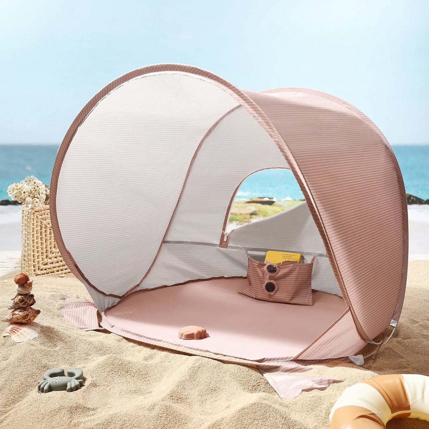 The Tiny Land® One-Stop Beach Toy & Tent Set – Blush Pink features a small pink and white tent with UV protection, a sand play set, and a woven basket, perfect for seaside fun under the clear blue sky.|Blush Pink