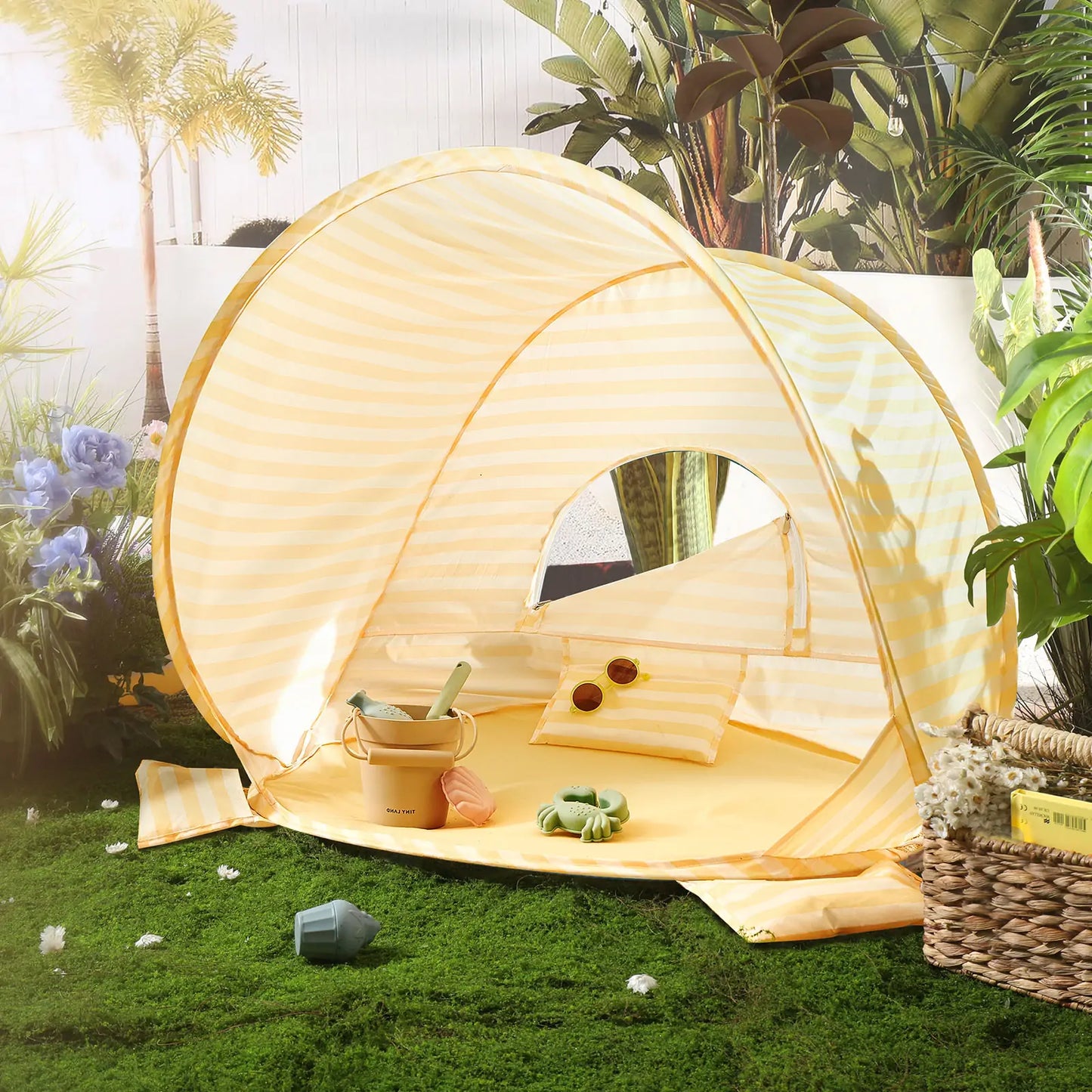 The Tiny Land® One-Stop Beach Toy & Tent Set – Pastel Yellow Stripes features a compact striped tent on artificial grass, with a pillow, sunglasses, pillow roll, and a pastel sand play set bucket surrounded by potted plants and flowers.|Pastel Yellow Stripes