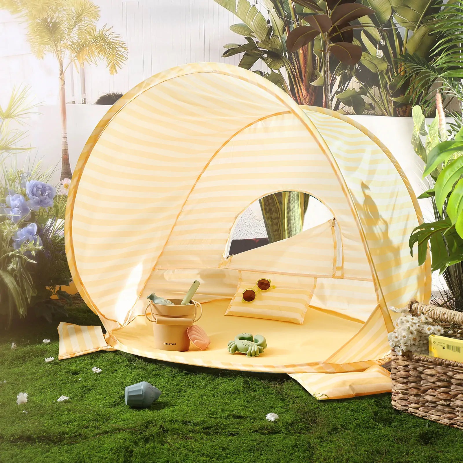The Tiny Land® One-Stop Beach Toy & Tent Set – Pastel Yellow Stripes features a compact striped tent on artificial grass, with a pillow, sunglasses, pillow roll, and a pastel sand play set bucket surrounded by potted plants and flowers.|Pastel Yellow Stripes