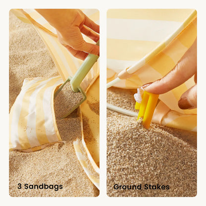 A hand fills a sandbag for the Tiny Land® One-Stop Beach Toy & Tent Set – Pastel Yellow Stripes, while another secures a yellow ground stake beside the striped fabric. Text reads “3 Sandbags” and “Ground Stakes.”.|Pastel Yellow Stripes