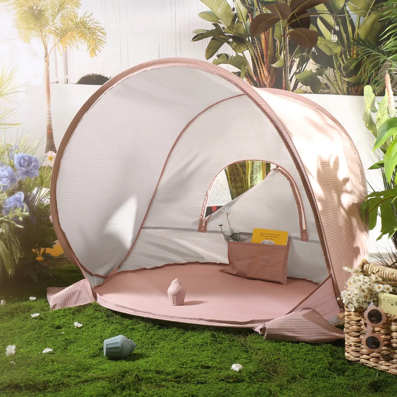 The Tiny Land® One-Stop Beach Toy & Tent Set – Blush Pink, a small UV-protected pink and white striped tent, is set up on grass among flowers, a basket, and plants with sunlight streaming in from the left.|Blush Pink