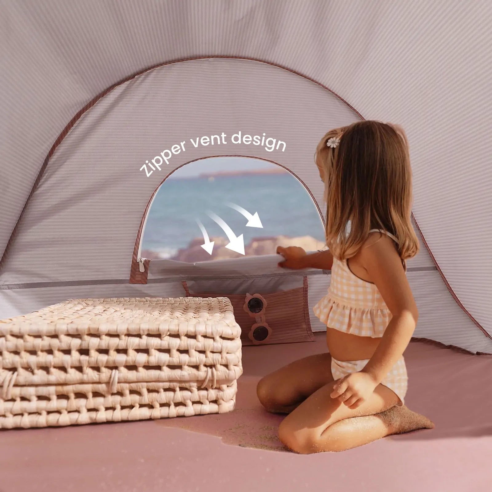 A young girl sits inside a Tiny Land® One-Stop Beach Toy & Tent Set – Blush Pink, gazing at the ocean. Woven mats are stacked nearby and sand covers the tent floor, highlighting this UV-protective set by Tiny Land.|Blush Pink