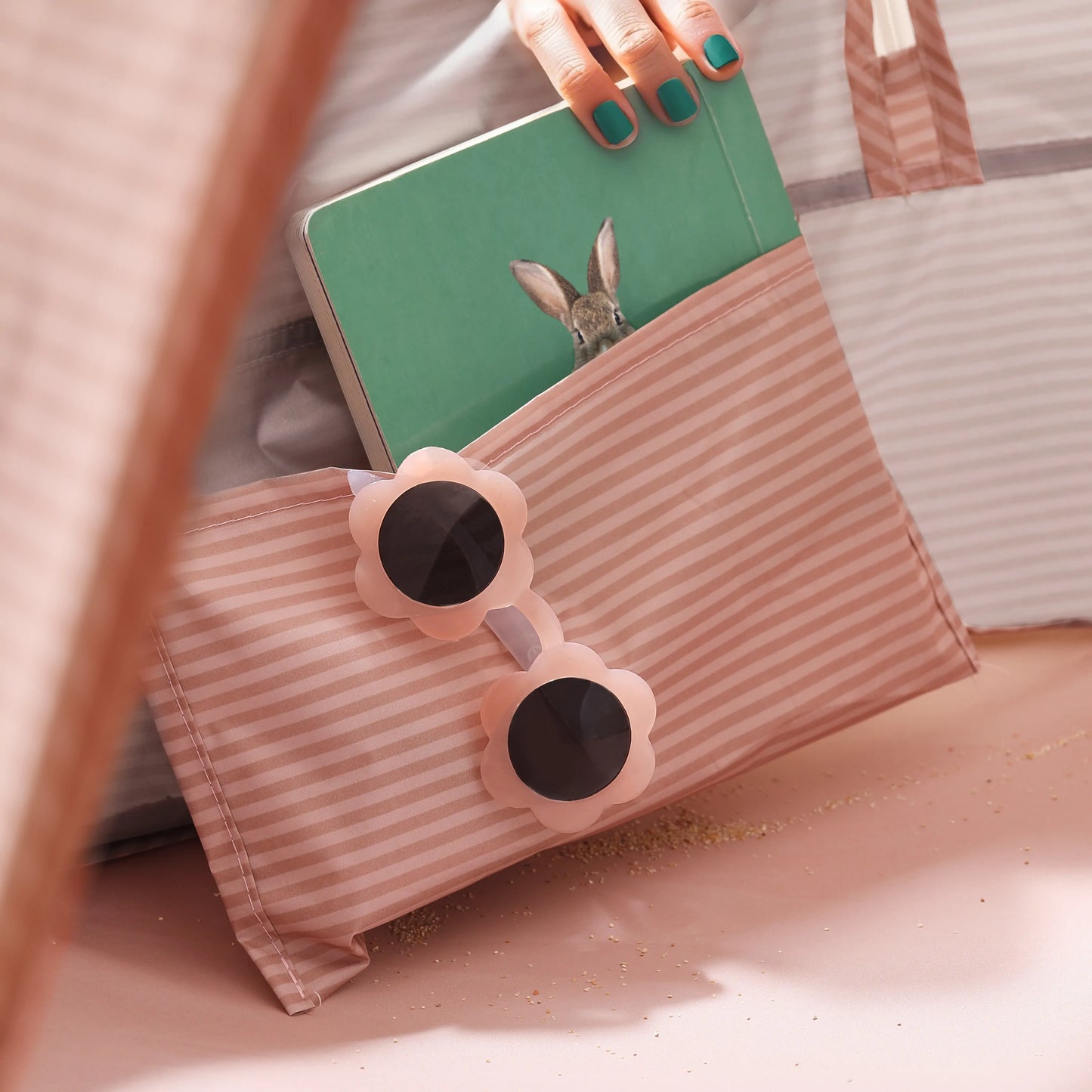 A hand with teal nail polish packs a green rabbit book into the Tiny Land® One-Stop Beach Toy & Tent Set – Blush Pink bag. Flower-shaped UV-protective sunglasses rest on top, all arranged on a pink surface.|Blush Pink