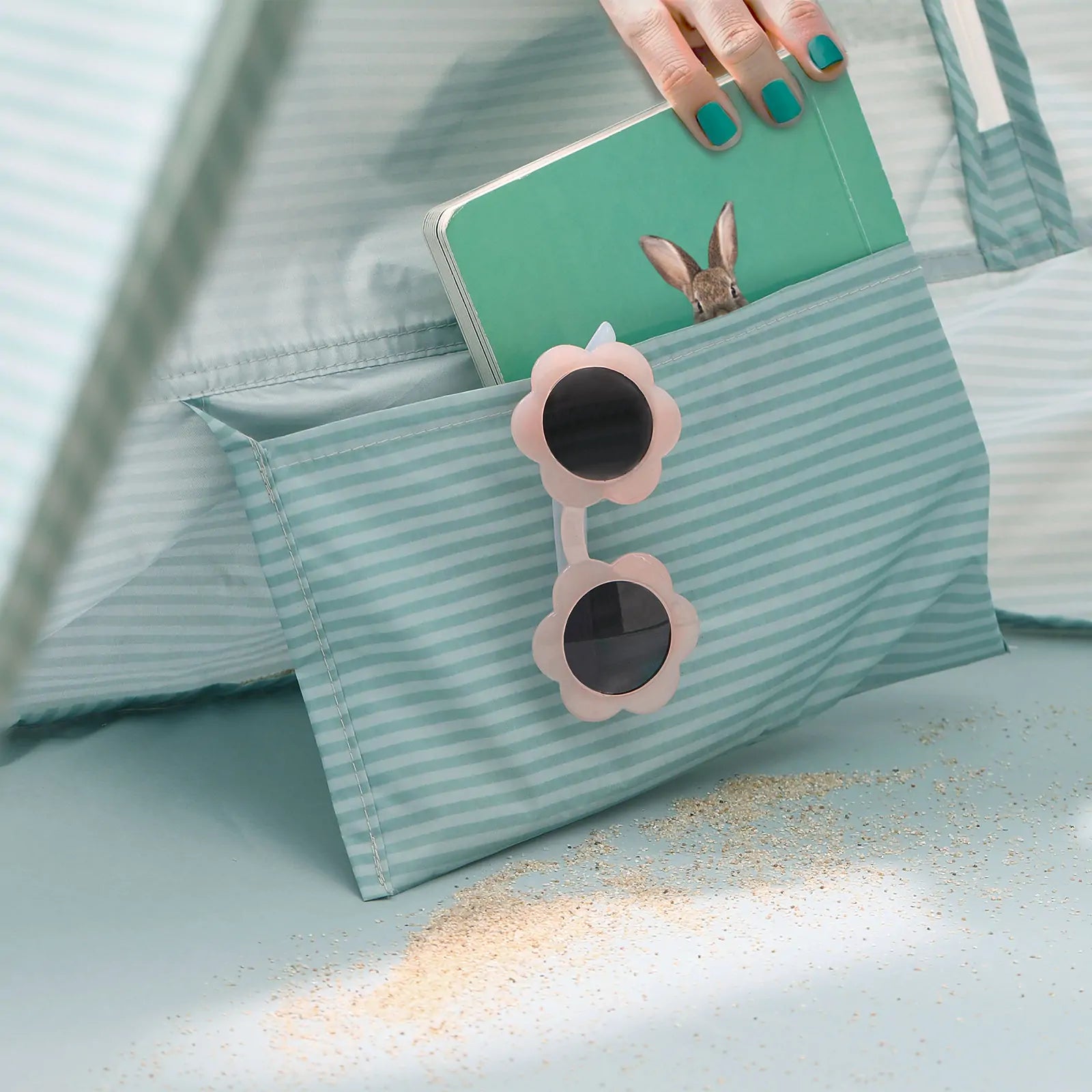 A hand with teal-painted nails holds a notebook near the Tiny Land® One-Stop Beach Toy & Tent Set – Sea Blue, with pink flower-shaped sunglasses and scattered sand adding to the relaxing beach vibe.|Sea Blue