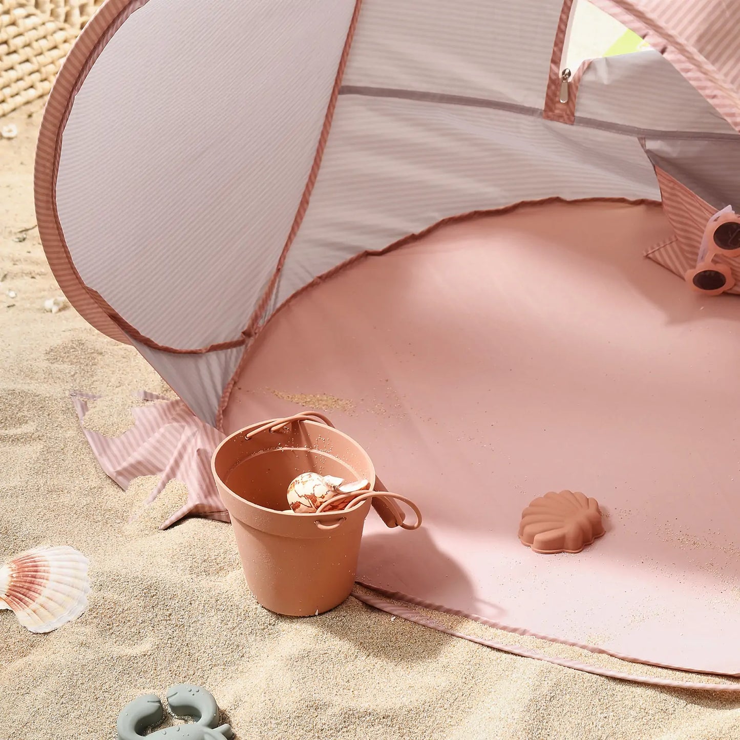 The Tiny Land® One-Stop Beach Toy & Tent Set – Blush Pink is set up on the sand, with a brown bucket holding a seashell and toy, a pink shell-shaped mold, and scattered seashells. Sunlight creates soft shadows inside the UV-protective tent.|Blush Pink