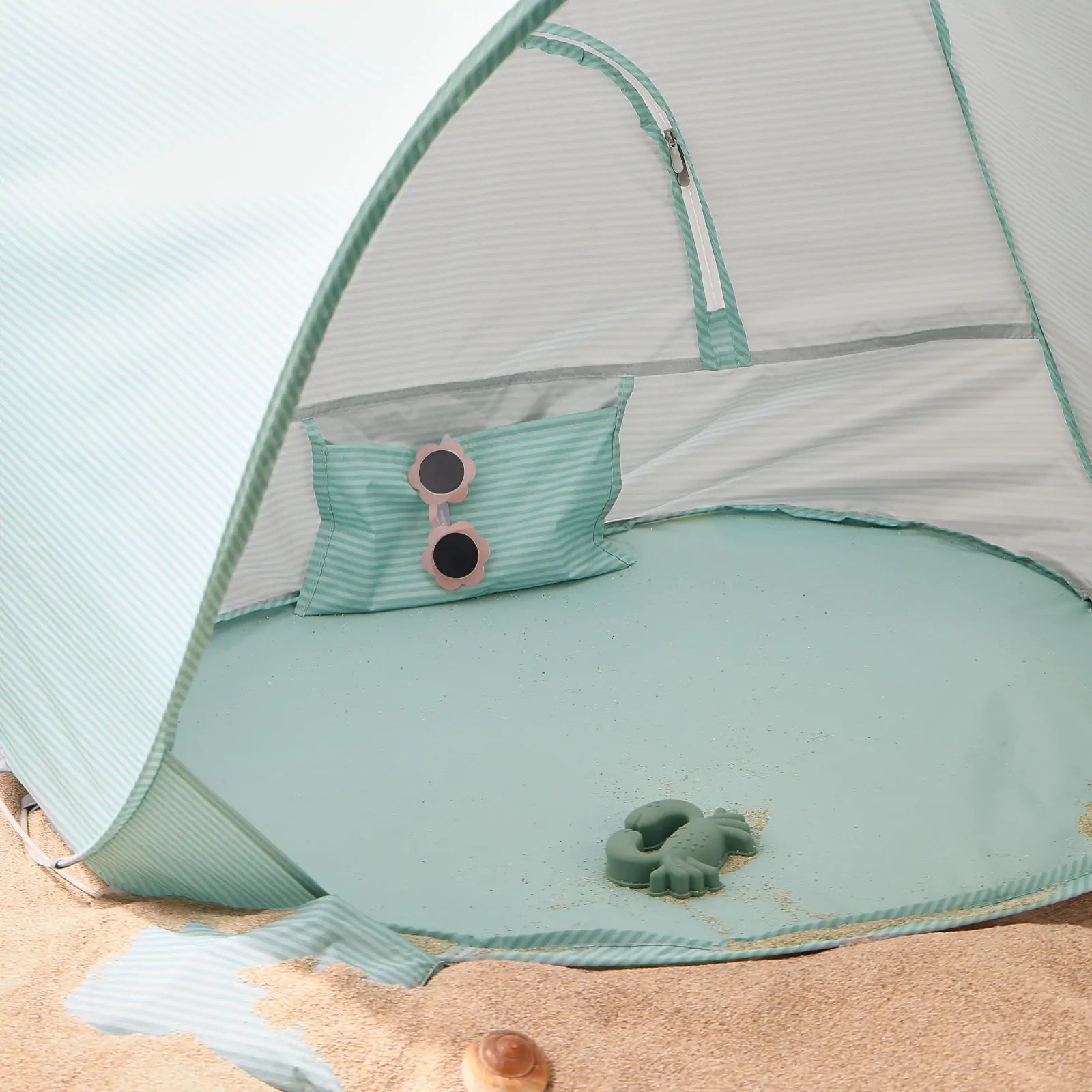 The Tiny Land® One-Stop Beach Toy & Tent Set – Sea Blue sits on the sand, complete with a matching pillow, round pink sunglasses, and a green toy turtle from Tiny Land. Some sand is scattered inside the UV-protective shade tent.|Sea Blue
