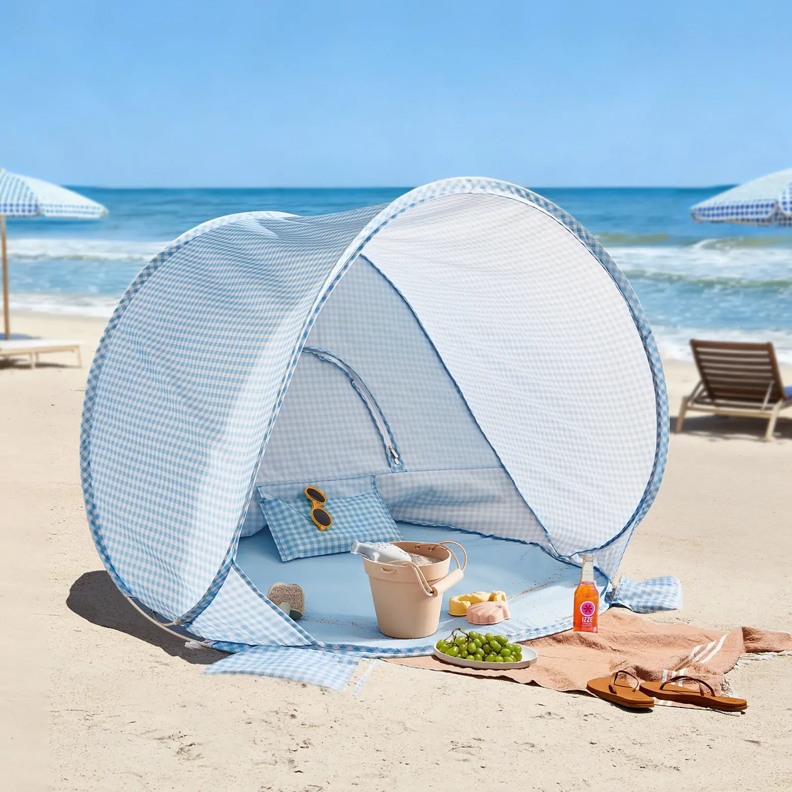 The Tiny Land® One-Stop Beach Toy & Tent Set in Light Blue Gingham sits on the sand near the ocean, with sunglasses and a beige bag inside. Nearby, a towel displays grapes, croissants, a drink, and sunscreen. Beach chairs and umbrellas are in the background.|Light Blue Gingham