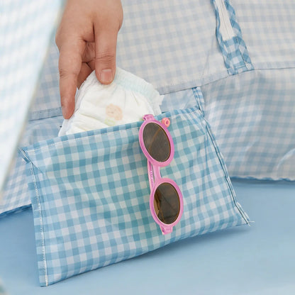 A hand puts a diaper into a blue gingham pouch with pink sunglasses, ideal for beach days with the Tiny Land® One-Stop Beach Toy & Tent Set – Light Blue Gingham. The matching pattern appears in the background.|Light Blue Gingham