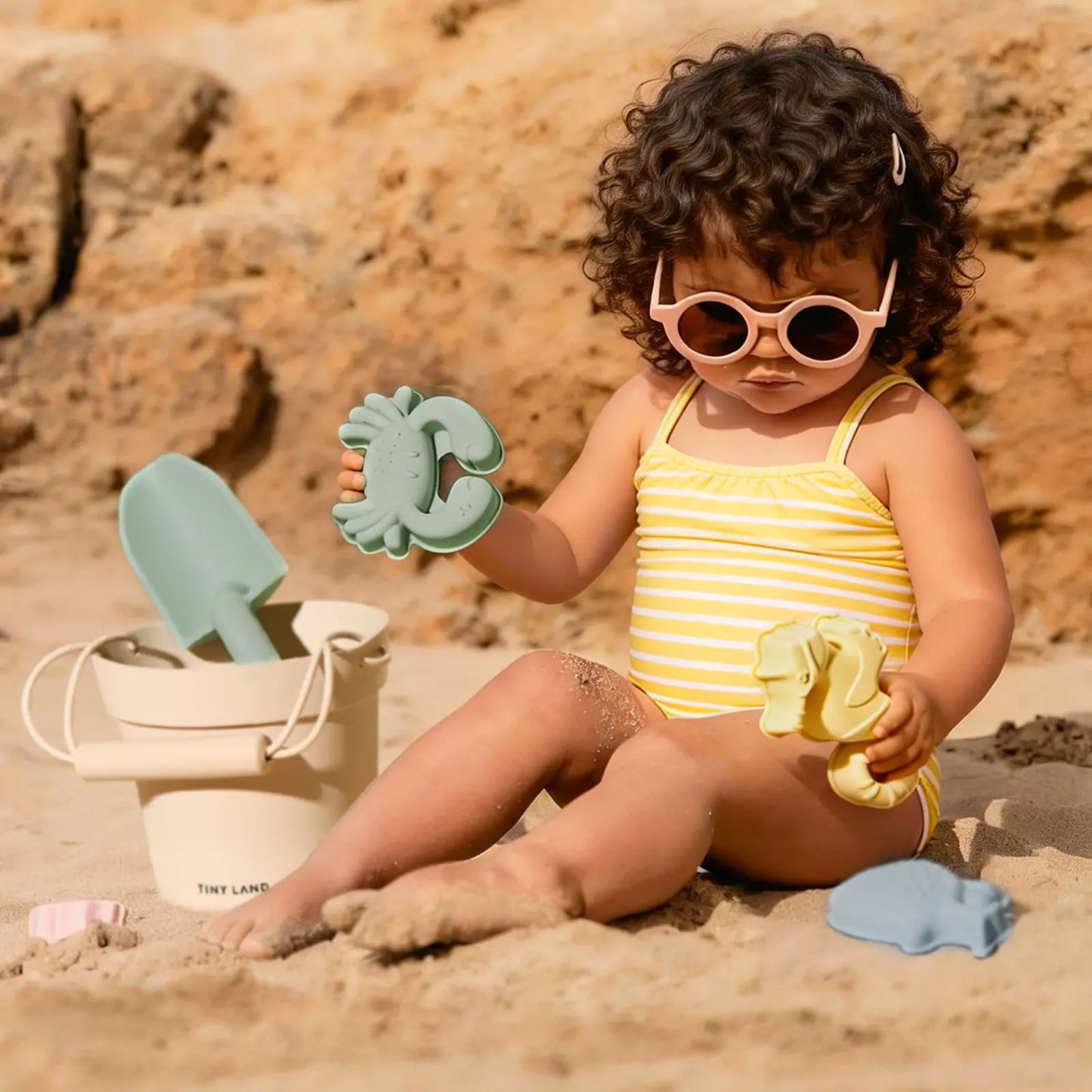 A toddler wearing a pastel yellow striped swimsuit and pink sunglasses plays on the sand with the Tiny Land® One-Stop Beach Toy & Tent Set, featuring a beige bucket and green shovel, with rocks in the background.|Pastel Yellow Stripes