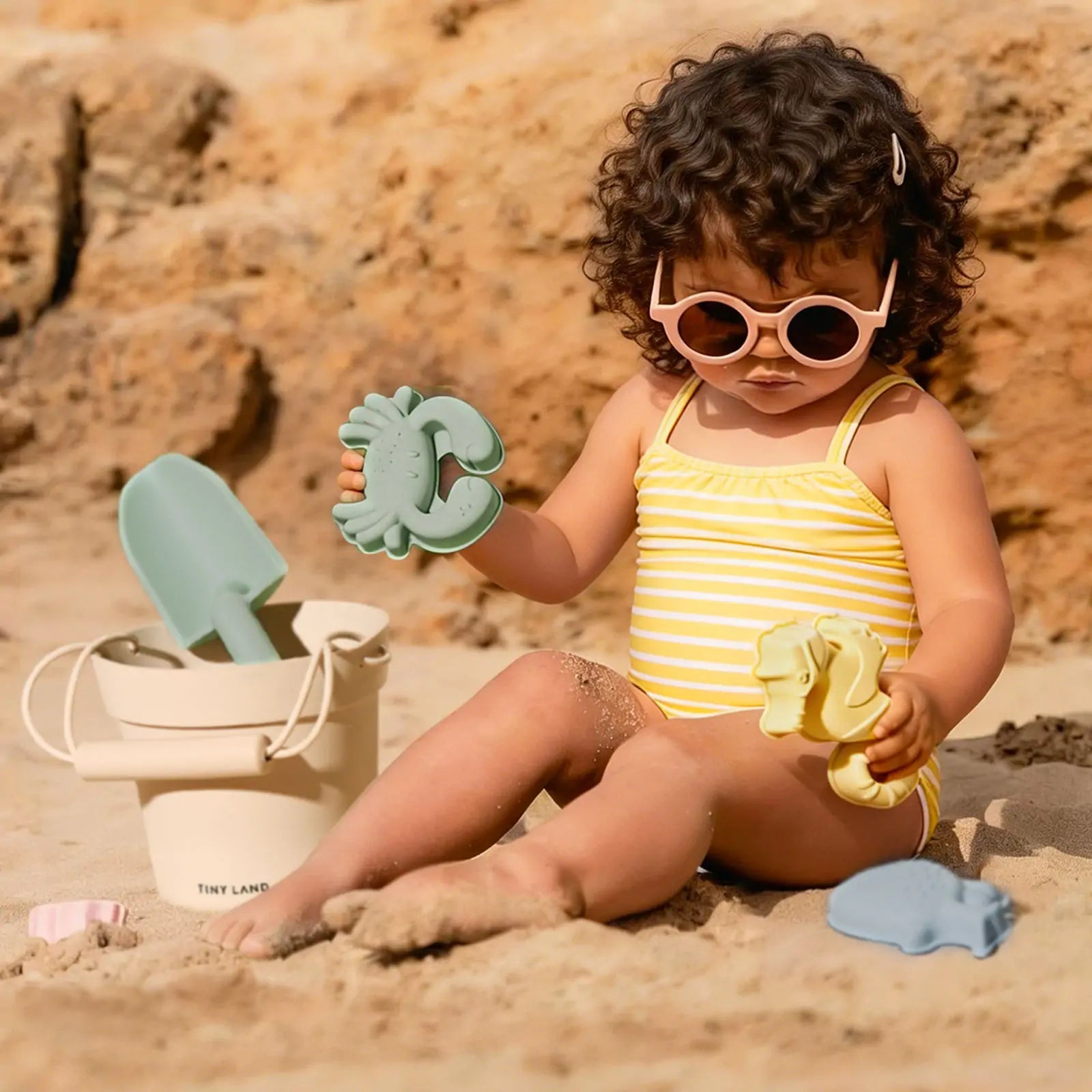 A toddler wearing a pastel yellow striped swimsuit and pink sunglasses plays on the sand with the Tiny Land® One-Stop Beach Toy & Tent Set, featuring a beige bucket and green shovel, with rocks in the background.|Pastel Yellow Stripes